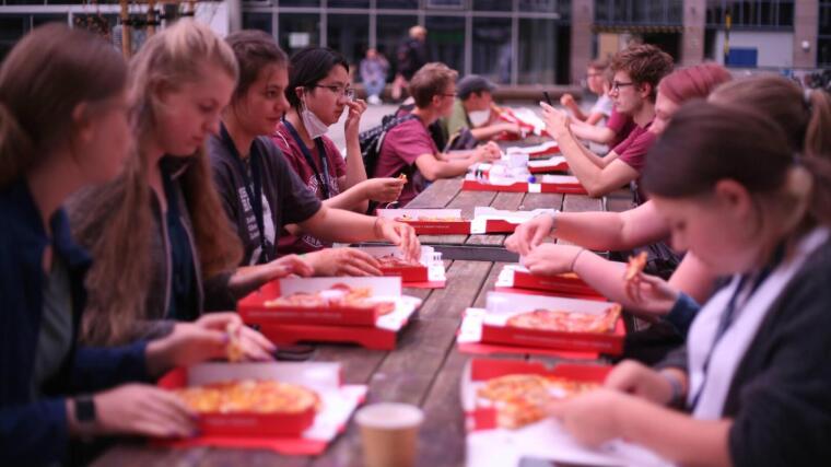Sommercamp-Teilnehmende sitzen gemeinsam draußen und essen Pizza