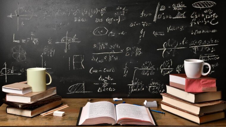 A wooden table in front of a blackboard. On the desk are books, chalk and coffee mugs.