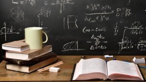 Ein Holztisch vor einer Kreidetafel. Auf den Tisch stehen Bücher, Kreide und eine Kaffeetasse.