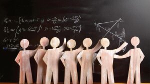 Small wooden figures with raised arms stand in front of a blackboard.