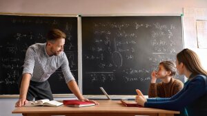 A teacher leans on a table at which students are sitting and is having a conversation.
