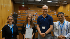 Nils Dyck und Juliane Sperling wird der Best Paper Award überreicht. Von links: Jacqueline Staub (Conference Chair), Juliane Sperling, Nils Dyck, Adish Singla (Conference Chair).