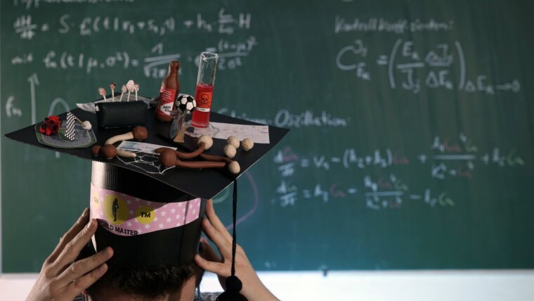 Wissenschaftler mit selbstgebasteltem Doktorhut vor einer beschriebenen Tafel